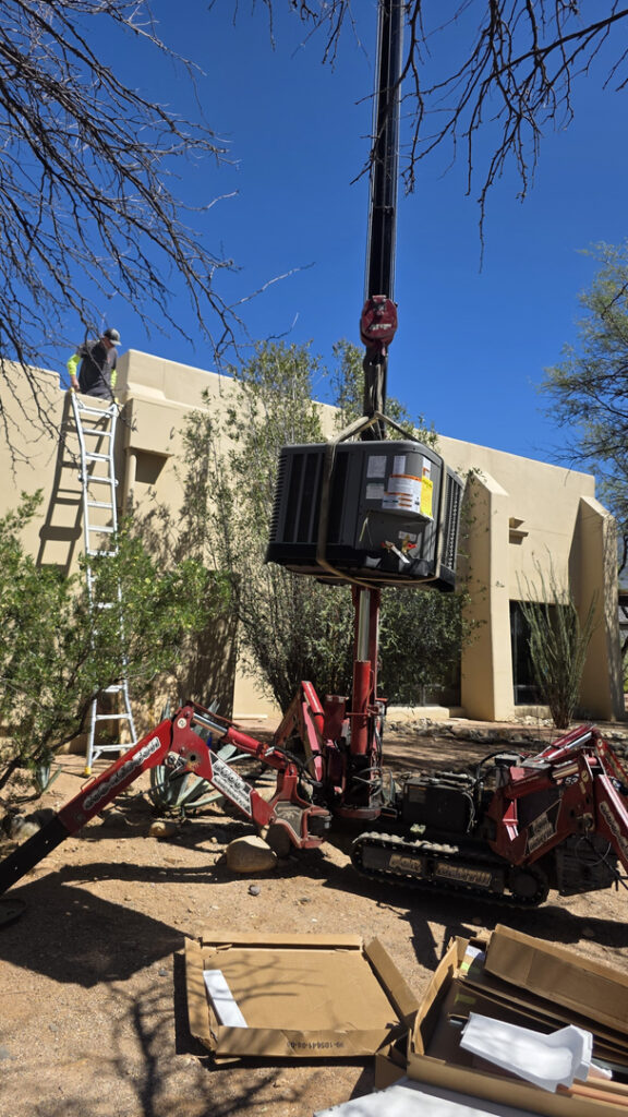 A technician carefully guides a new outdoor HVAC unit into place beside a Tucson home while a compact lifting machine hoists it overhead. Another technician works from a ladder near the roofline, with desert landscaping, bright blue skies, and a stucco exterior in the background. Professional HVAC installation helps keep Tucson homes comfortable through the heat.