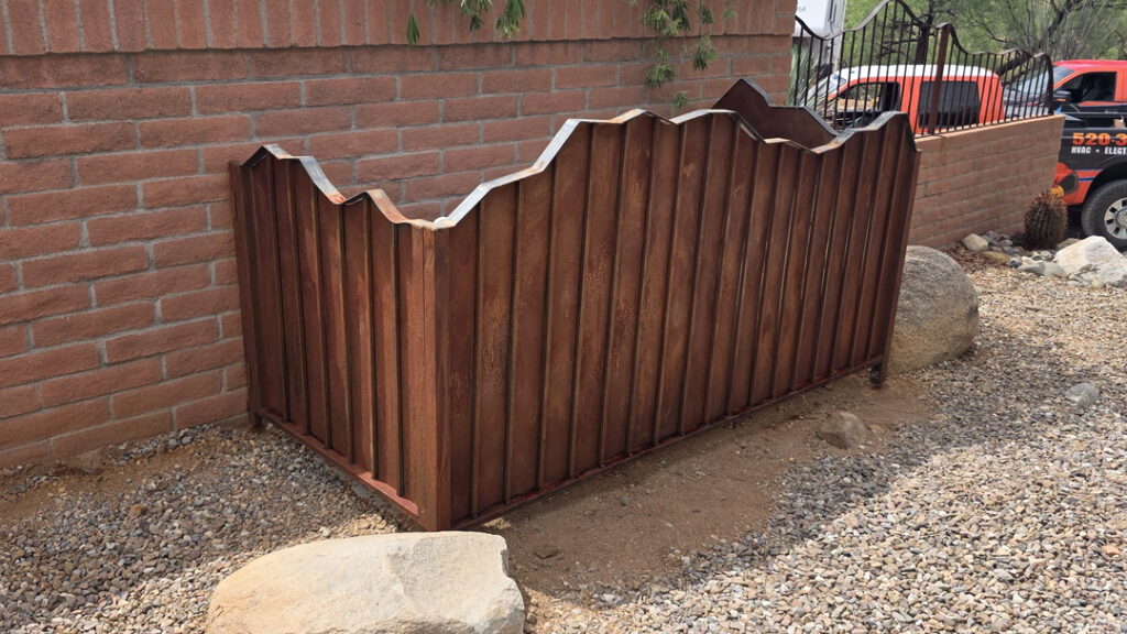 A weathered, rust-colored metal enclosure with a decorative wave-shaped top sits beside a brick wall in a Tucson gravel yard, helping conceal outdoor HVAC equipment. Desert landscaping, large rocks, and a service truck are visible in the background.