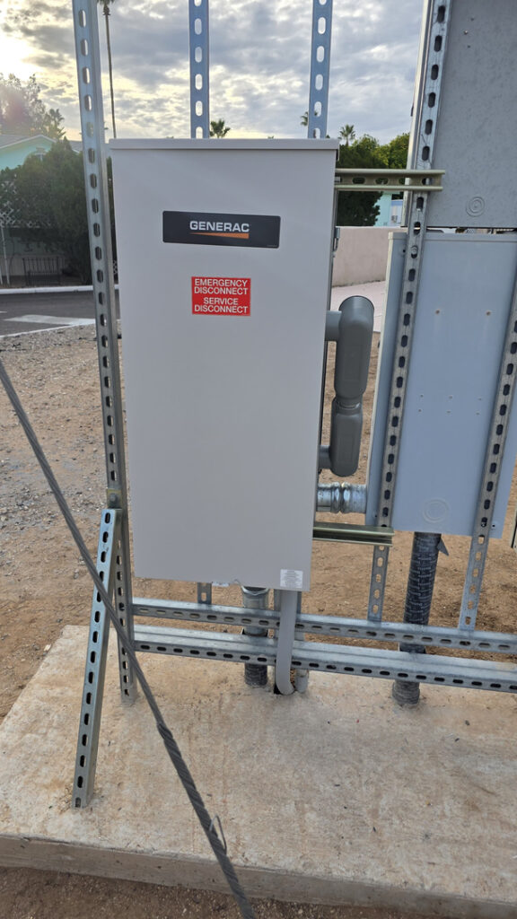 A close-up of a gray Generac emergency disconnect enclosure mounted on a metal support frame outdoors in Tucson. Red and black labels clearly identify the emergency and service disconnects. Conduit and wiring connect the enclosure to nearby electrical equipment, with a residential neighborhood, palm trees, and a partly cloudy sky in the background.
