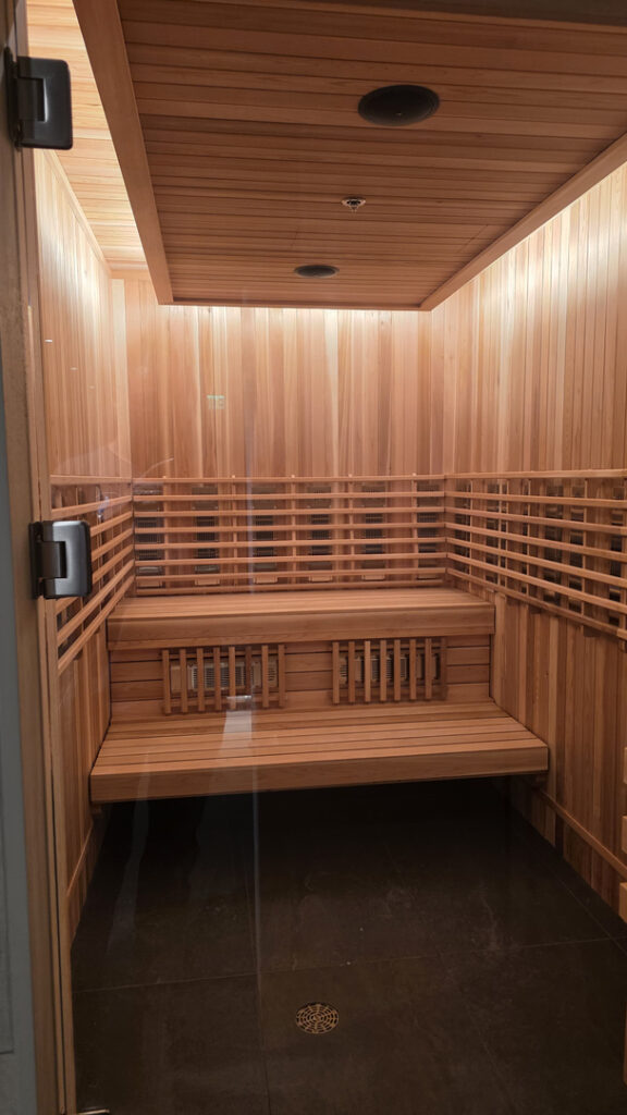 A warm, wood-paneled sauna in Tucson features tiered benches, horizontal safety rails, built-in lower vents, recessed ceiling fixtures, and dark tiled flooring with a central drainan inviting example of how thoughtful indoor comfort and ventilation design can elevate a space.