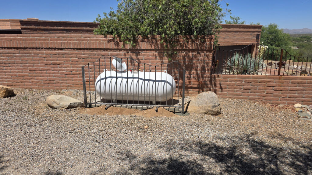 A white horizontal propane tank sits behind a black metal safety enclosure beside a reddish brick wall. Desert gravel, large rocks, agave, and a leafy tree surround the area, with Tucsons bright blue sky and distant mountain landscape in the background. Properly maintained outdoor equipment supports safe, dependable comfort for local Tucson homes.